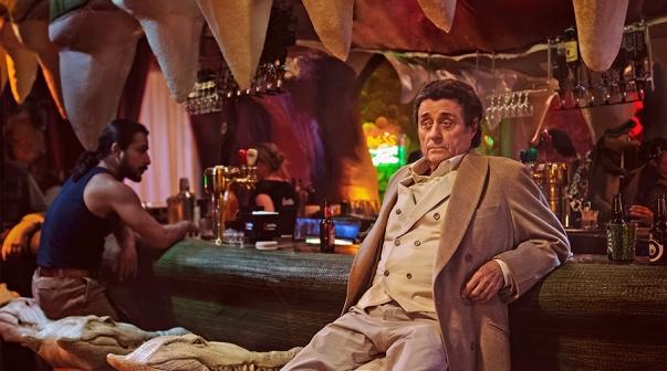 Ian McShane als Mr. Wednesday sitzt an einer Krokodilkopf-Bar