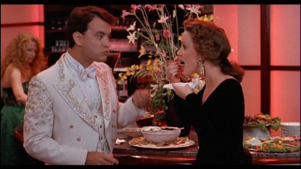 Tom Hanks in weißem Glitzeranzug streitet sich mit Frau vor Buffet